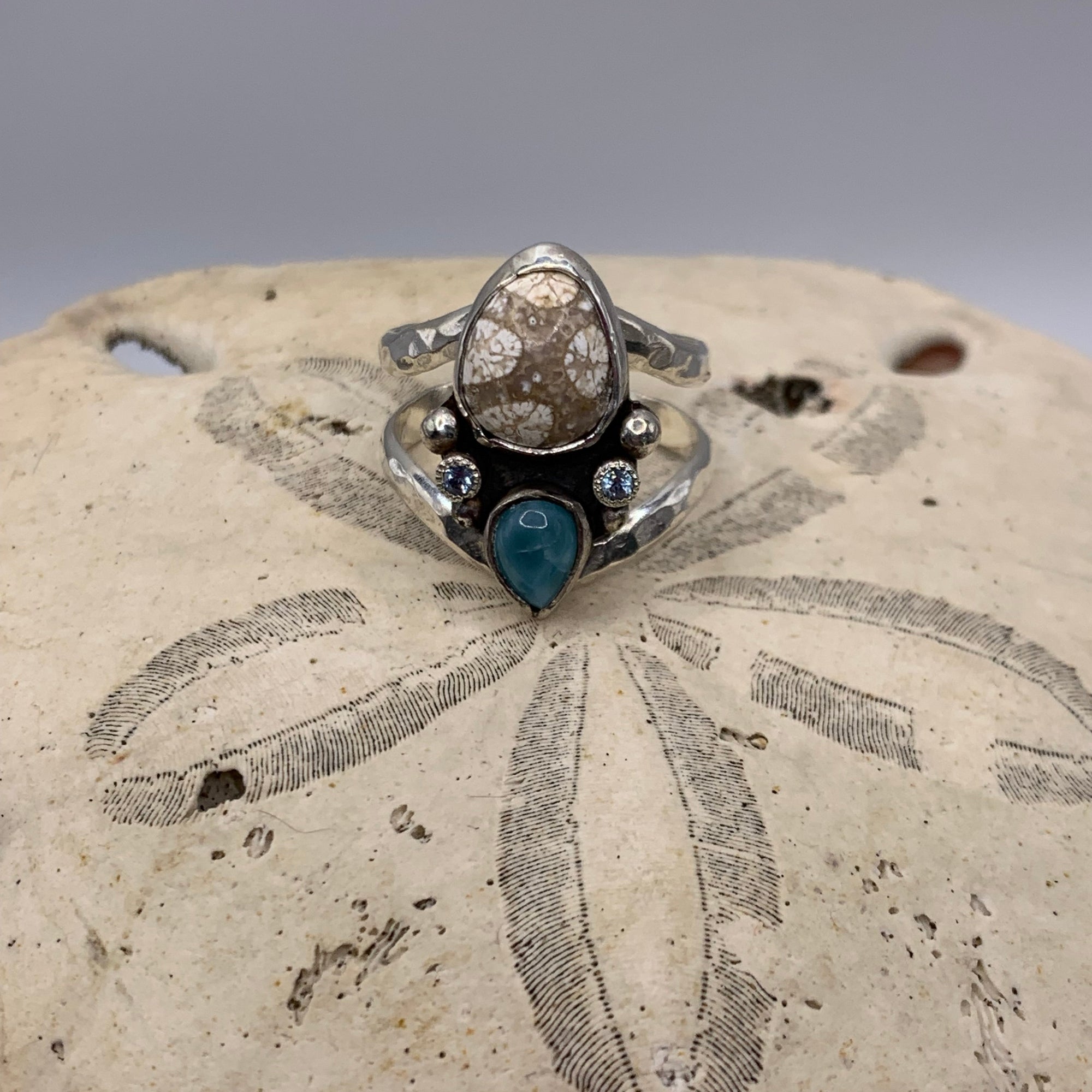 A sterling silver ring with a natural brown daisy stone and a teardrop-shaped turquoise accent, displayed on a textured sand dollar.