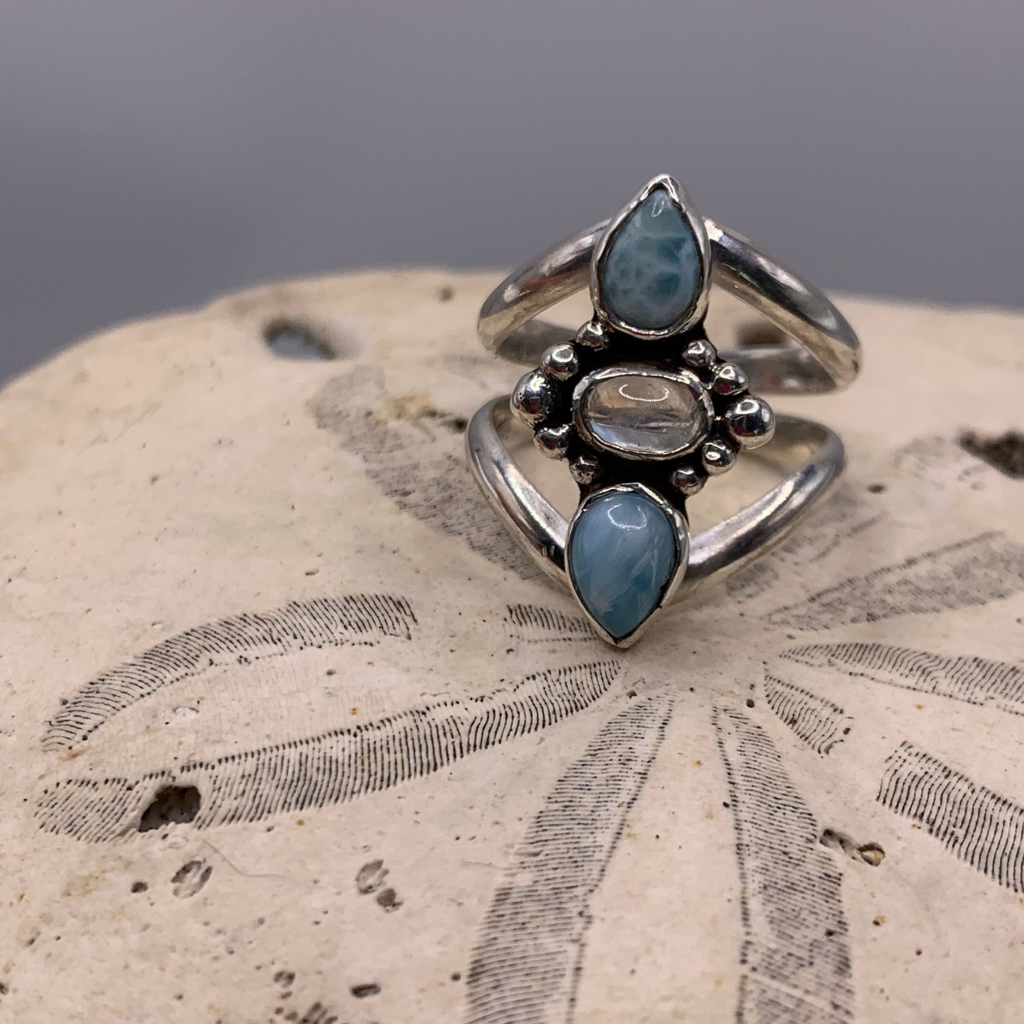 A silver ring with three light blue marbled gemstones, displayed on a textured beige stone surface against a neutral gray background.