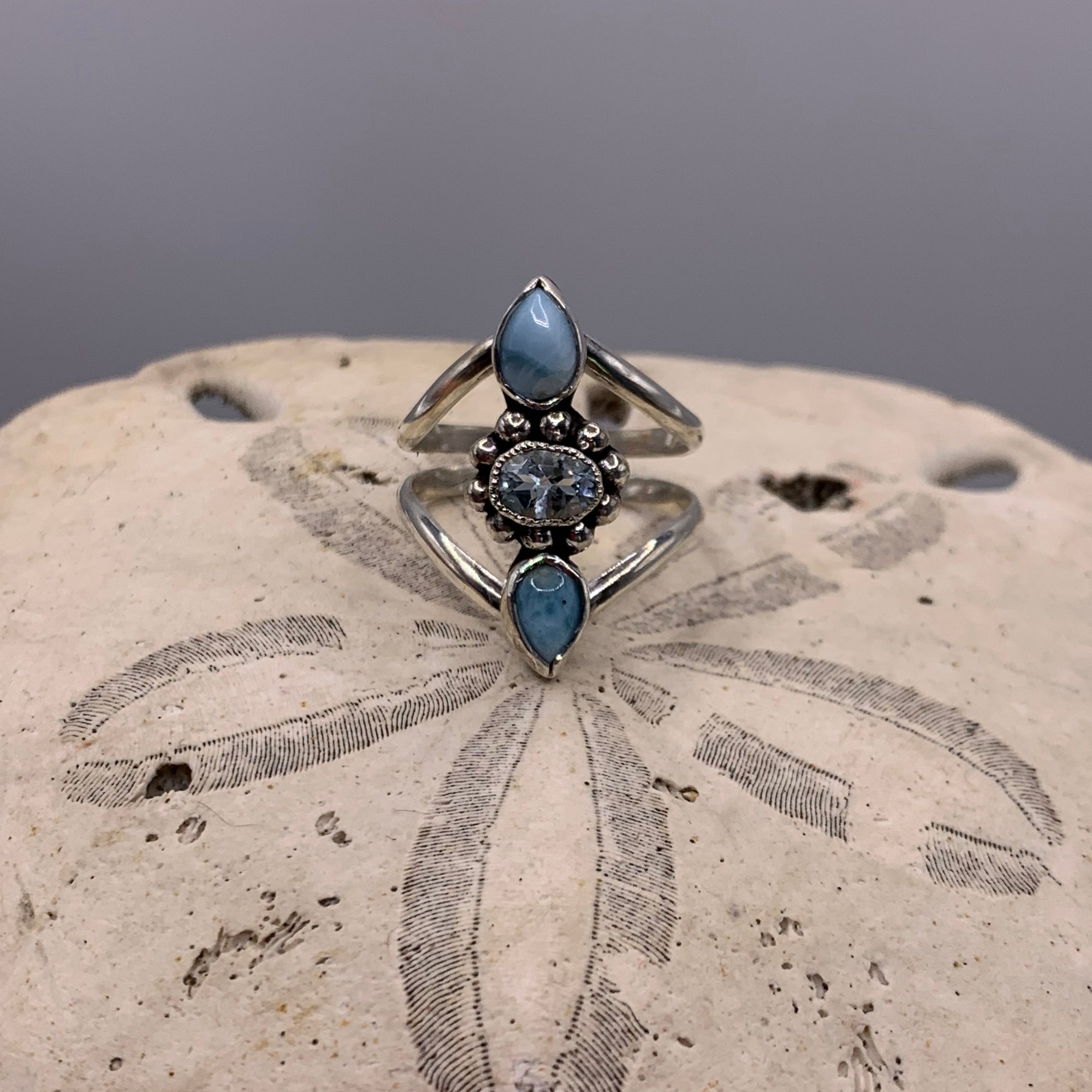 A silver ring with three marquise-cut light blue gemstones and a central round blue stone, displayed on a textured beige stone surface against a gray background.