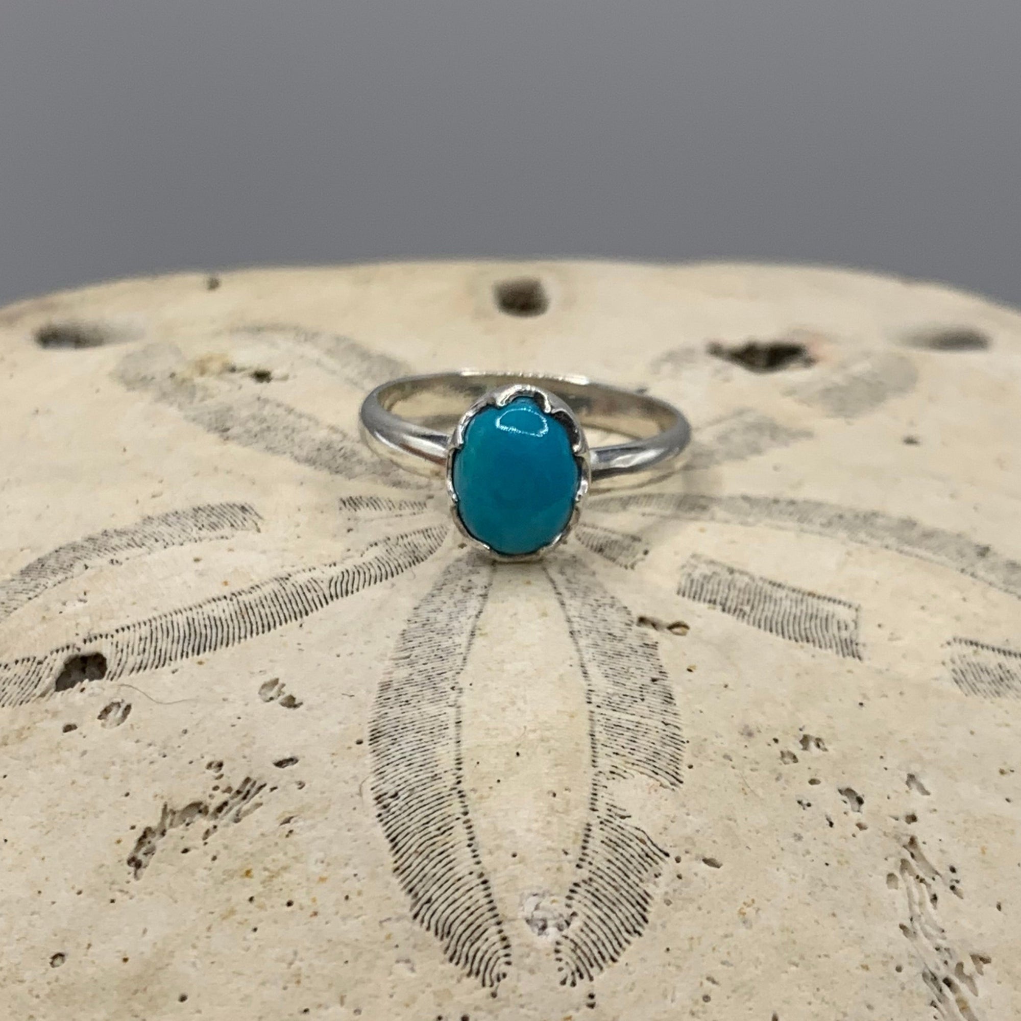 A sterling silver ring with an oval turquoise cabochon gemstone, displayed on a sand dollar against a neutral gray background.