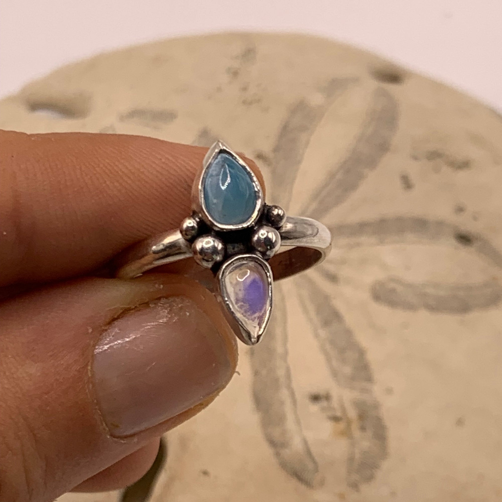 A hand holding a silver ring with two marquise-cut stones, one light blue and one iridescent lavender-purple, set against a sandy background.
