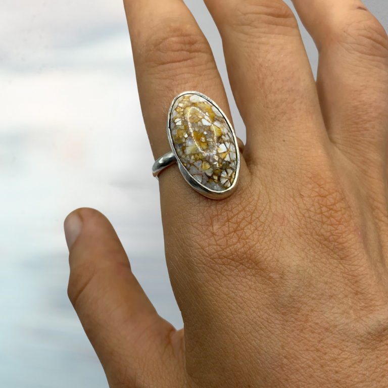 A close-up of a hand wearing a silver ring with a large, irregular yellow stone, set against a soft, out-of-focus light blue and white background.