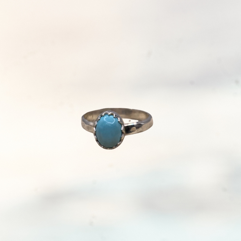 A silver ring with an oval light blue turquoise stone, shown worn on a person's finger against a blurred outdoor background.