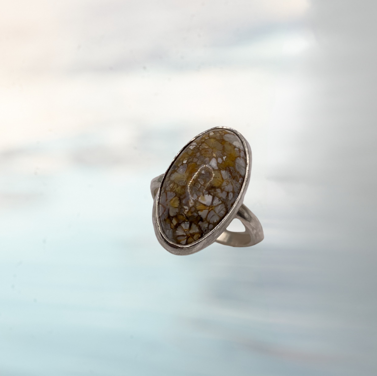 A silver ring featuring a large, oval-shaped cabochon stone filled with gold and white glitter, set against a soft, out-of-focus light blue and white background.