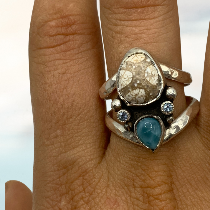 A silver ring with a large textured clear stone and a blue teardrop gemstone, shown worn on a hand against a blurred outdoor background.