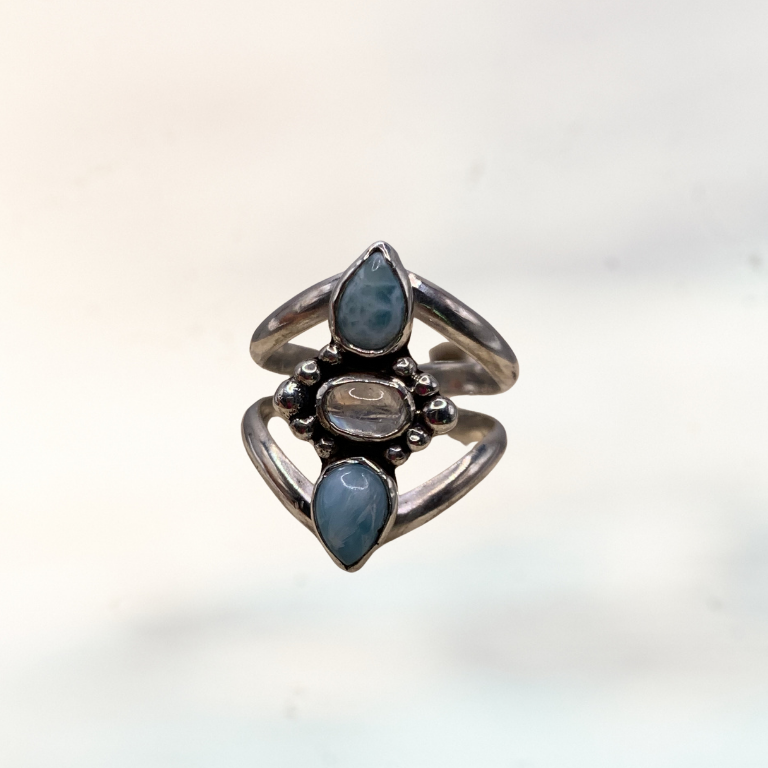 A close-up of a hand wearing a silver ring with three light blue gemstones and a central iridescent moonstone.