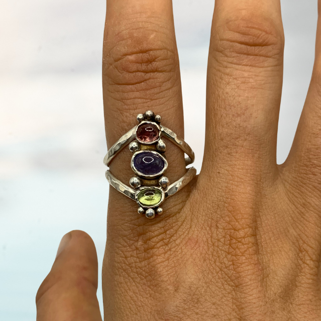 A close-up of a hand wearing a silver ring with three small oval gemstones in red, purple, and green.