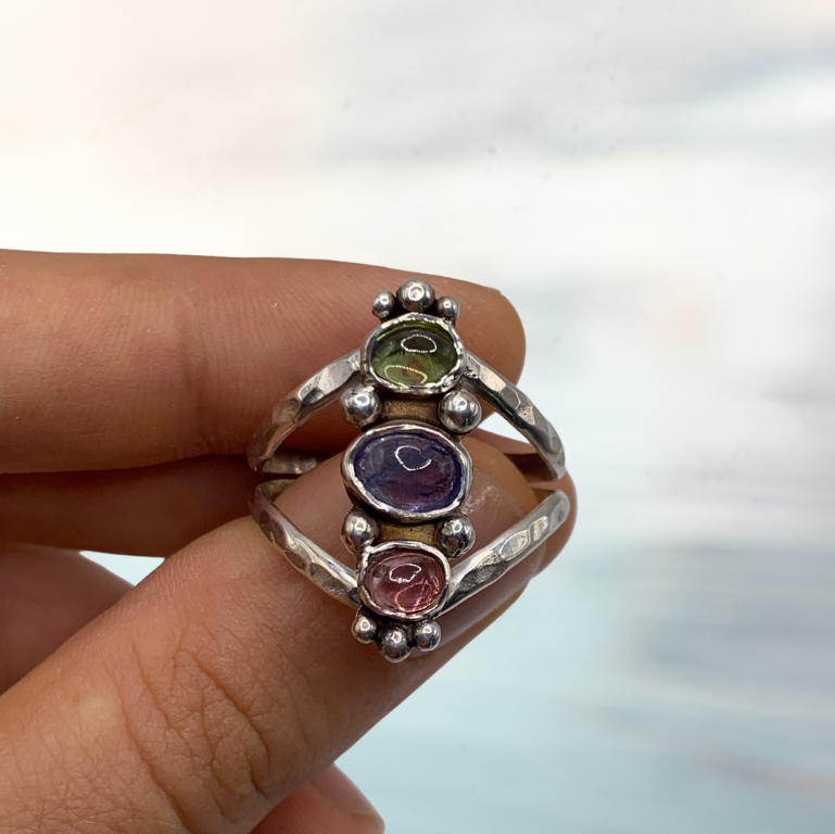 A hand holding a silver chakra ring with three colorful gemstones (green, purple, and pink) against a blurred light gray background.
