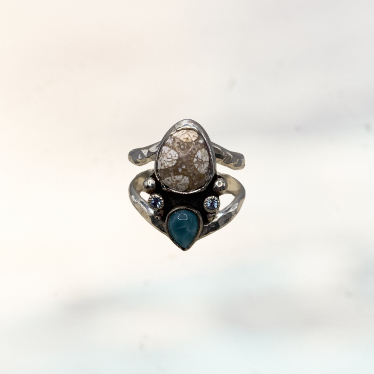 A silver ring featuring a large teardrop-shaped clear stone with gold inclusions, accented by a small blue teardrop gemstone and tiny clear stones, shown against a light, out-of-focus background.