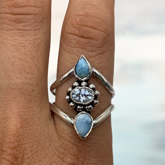 A close-up of a hand wearing a silver ring with three light blue teardrop-shaped stones and a central clear round stone, set against a blurred outdoor background.